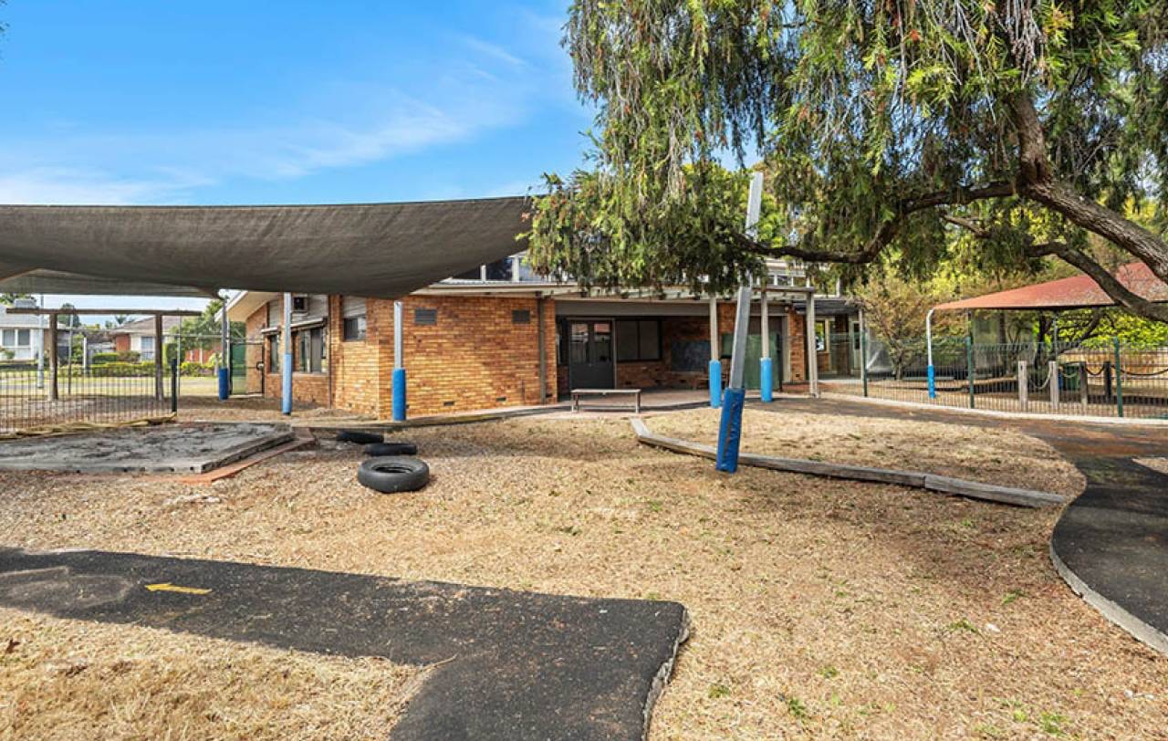 Rear of the brick building seen from the play yard, with shade sails and a mature peppercorn tree.