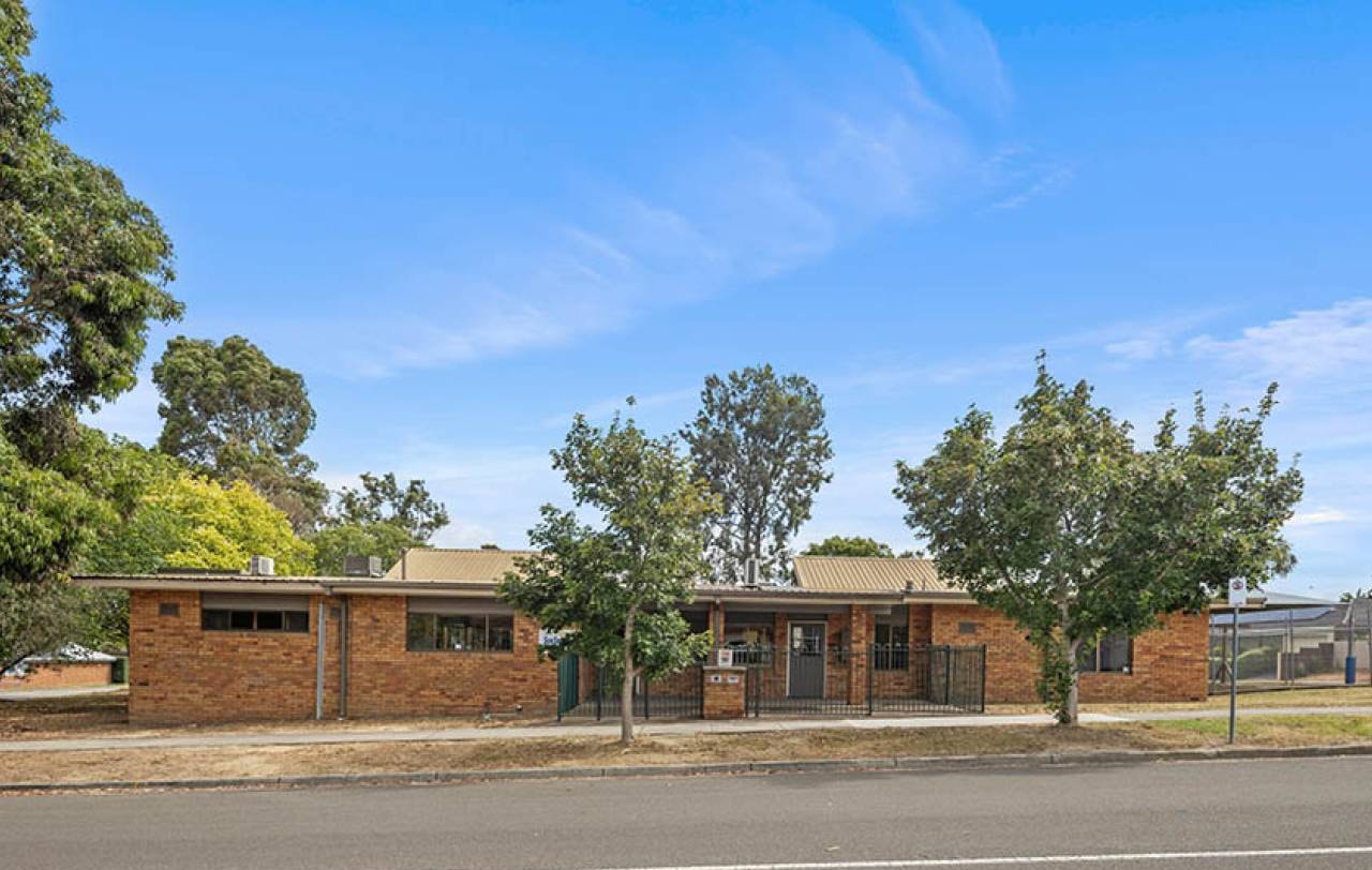 Single-storey brick building seen from the street, with mature trees along the frontage.